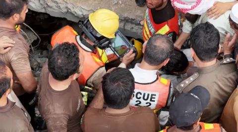 A group of men are seen from above standing around a man in high vis with a helmet on looking at a screen near a collapsed building