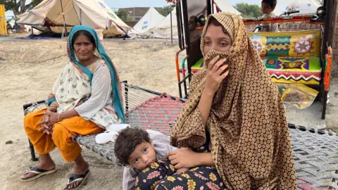 A woman drawing her headscarf across her face sits with a child and another woman wearing a headscarf